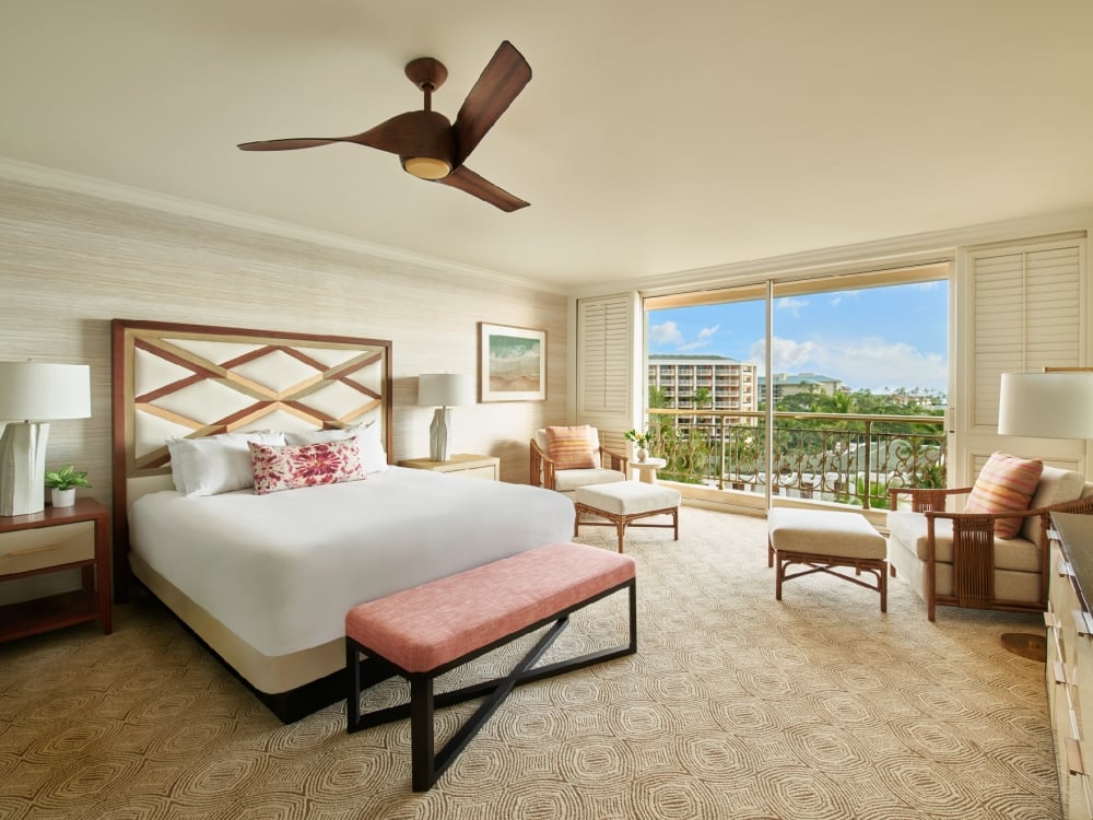 A resort guestroom featuring a large bed with a bench at the foot of it, two seating areas and an adjacent balcony