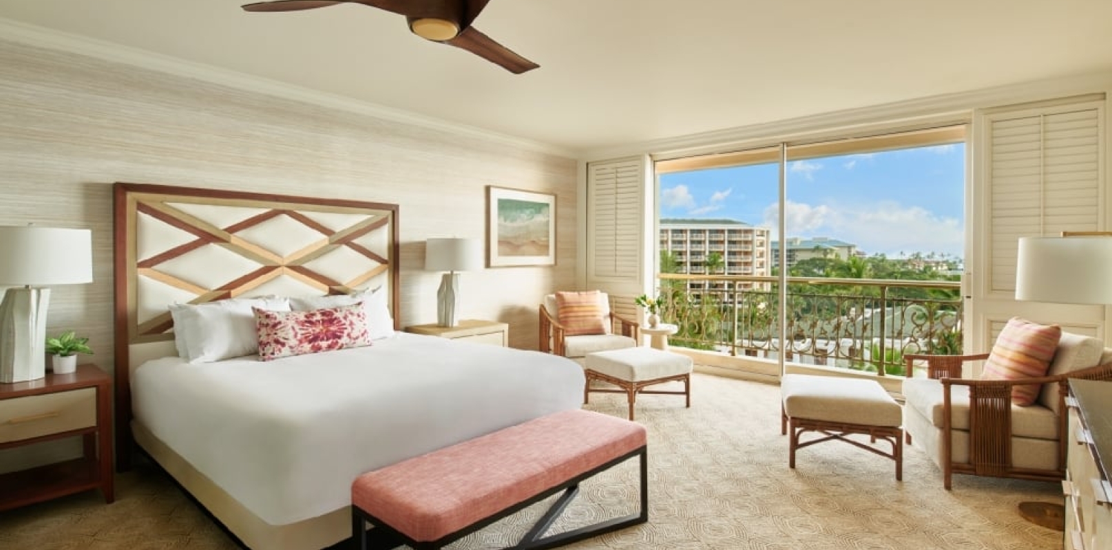 A resort guestroom featuring a large bed with a bench at the foot of it, two seating areas and an adjacent balcony